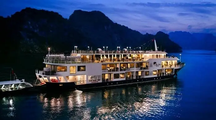 Luxury overnight cruise sailing through limestone islands in Ha Long Bay at sunset with calm emerald waters.