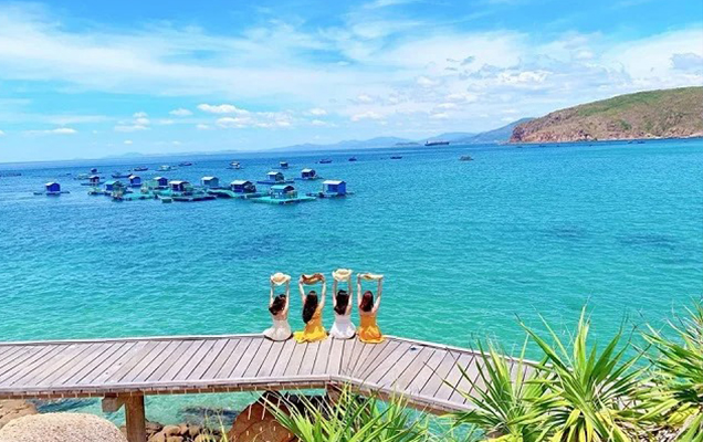 Four travelers sitting on a wooden deck overlooking the turquoise sea, floating houses, blue sky, and coastal hills in Quy Nhon, Vietnam.