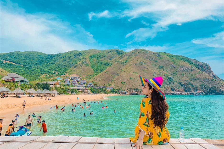 A woman sitting by the turquoise sea at Ky Co Beach in Quy Nhon, Vietnam, with sandy beach, green mountains, and clear blue sky.