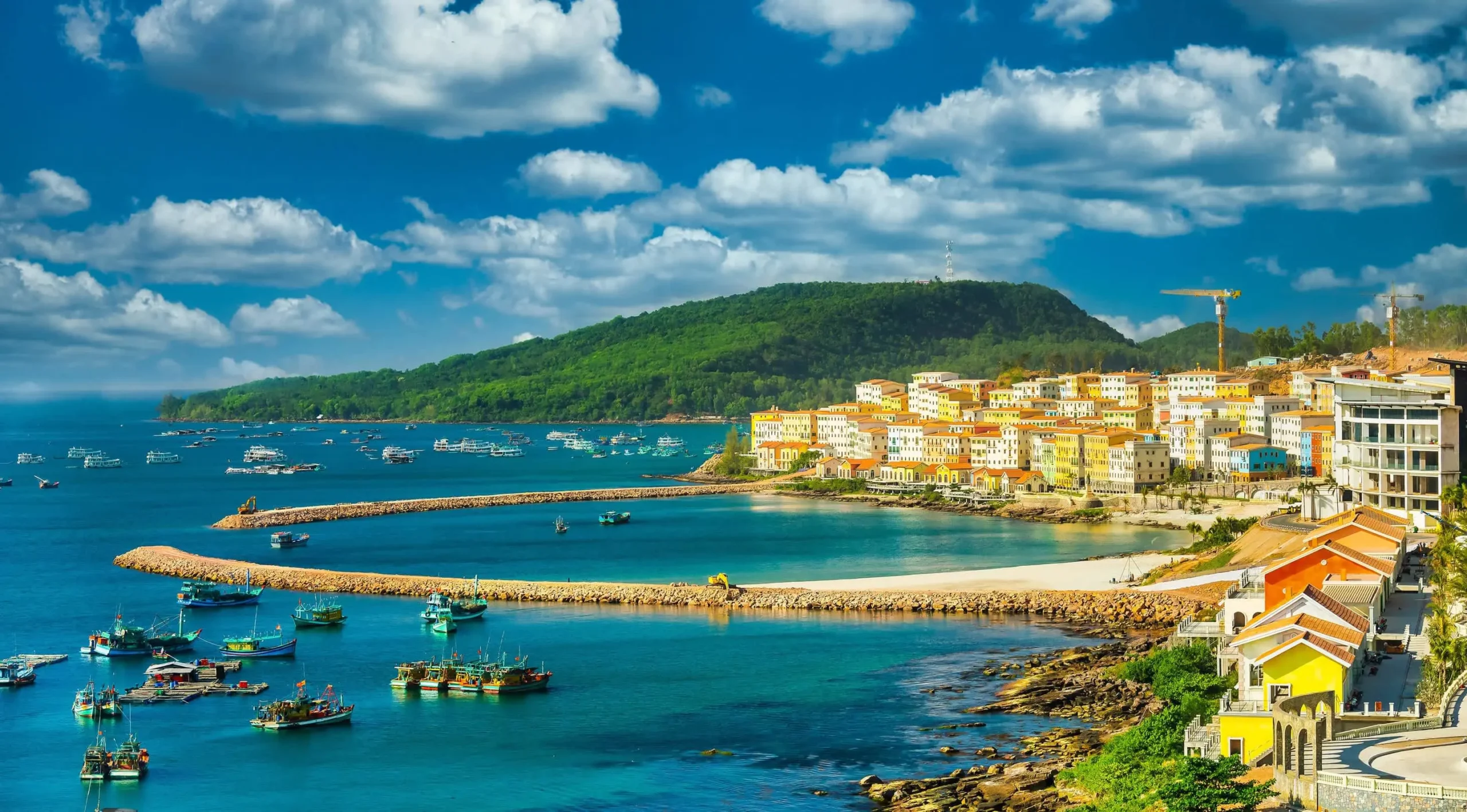 A panoramic coastal view in Phu Quoc Vietnam featuring Mediterranean-style architecture, turquoise waters, fishing boats, sandy beaches, and lush green hills under a bright blue sky.