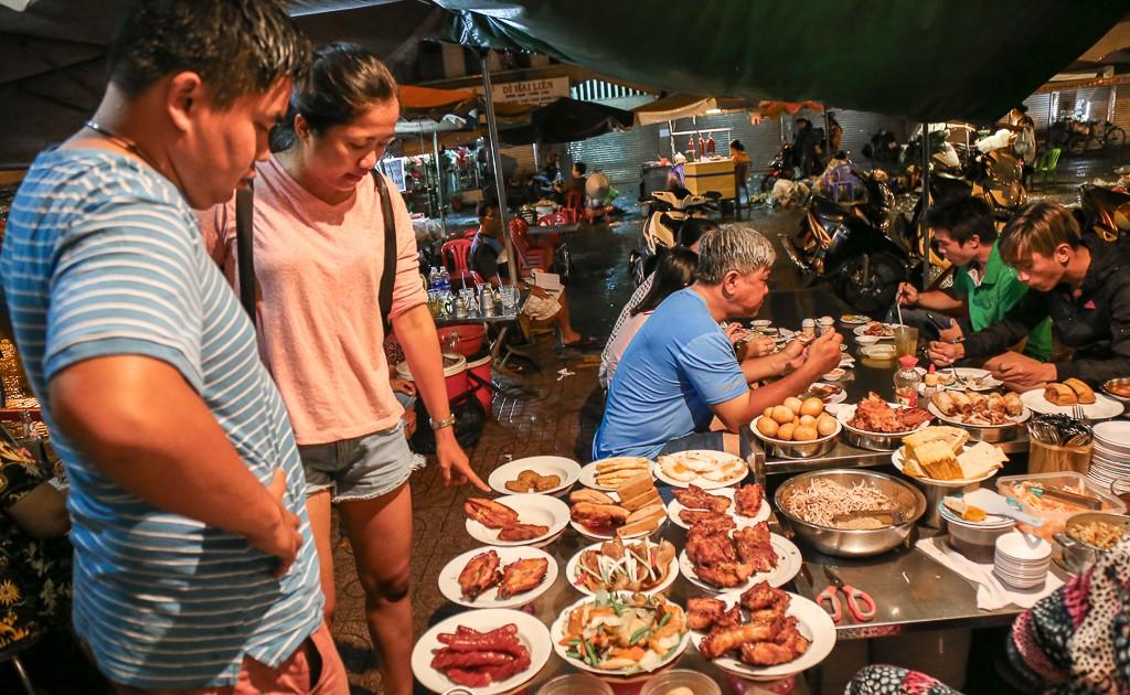 Locals and tourists choosing dishes at a bustling nighttime street food stall in Ho Chi Minh City, featuring popular Vietnamese dishes like Com Tam ingredients, sausages, and grilled meats.