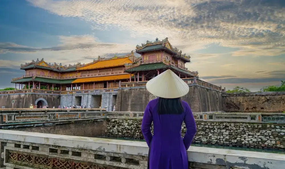 A woman in a traditional purple Ao Dai and conical hat gazing at a magnificent ancient palace gate across a stone moat.